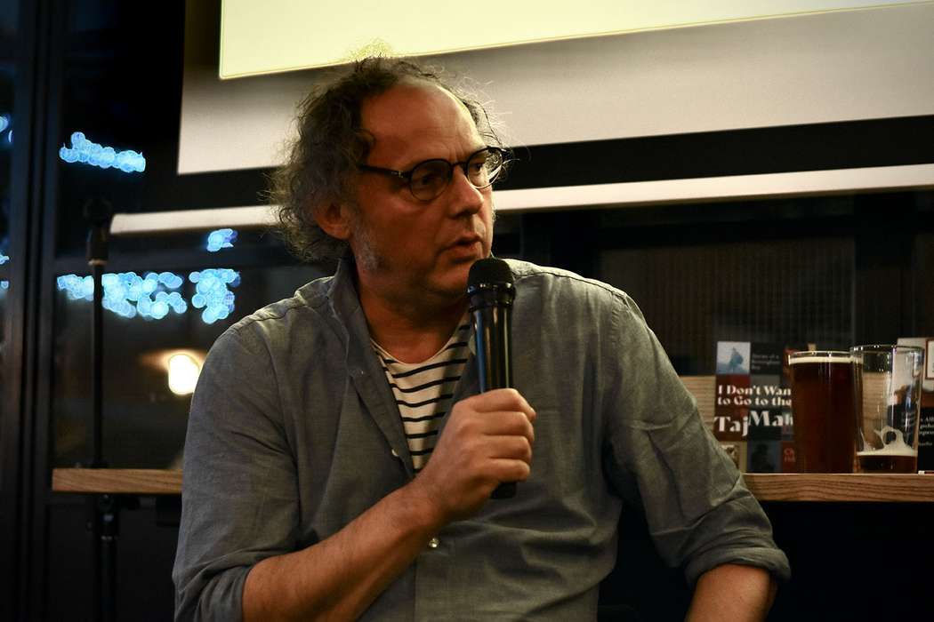Writer Charlie Hill holding a microphone at an indoor event with his book I Don't Want to go to the Taj Mahal in the background